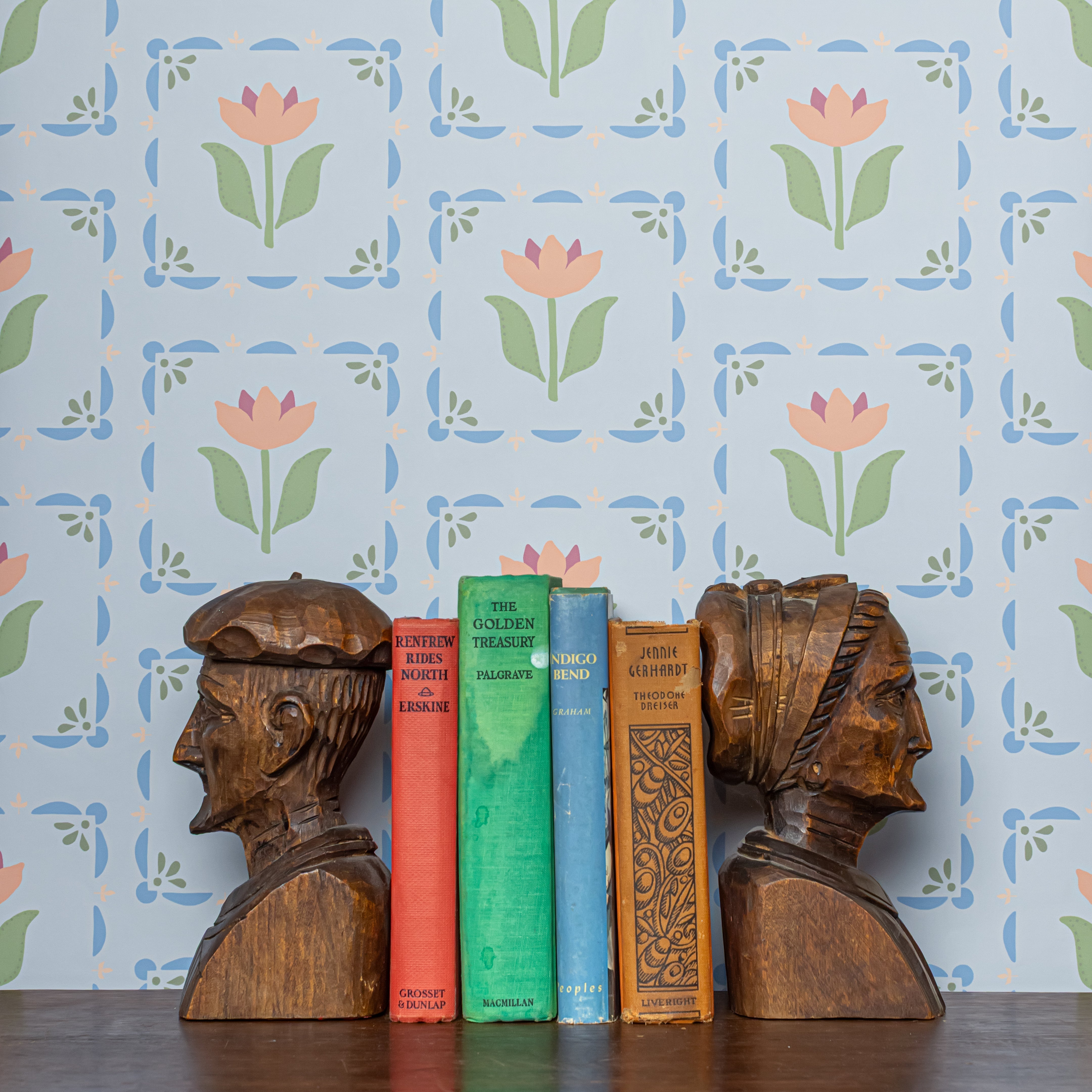 Wooden bookends shaped like heads on a shelf with books against a floral wallpaper background