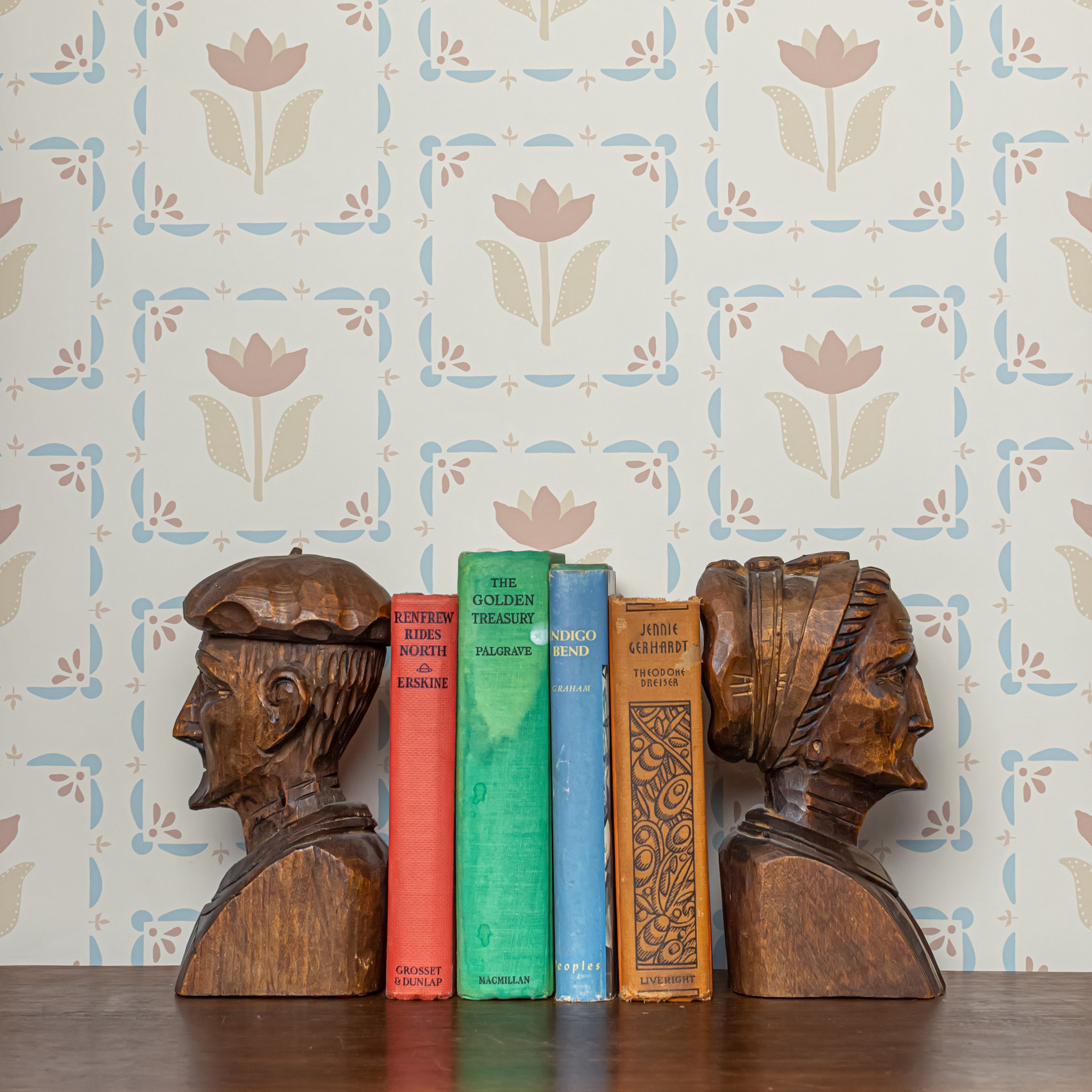 Wooden bookends shaped like faces with books of different colors against a floral-taupe and blue patterned wall.