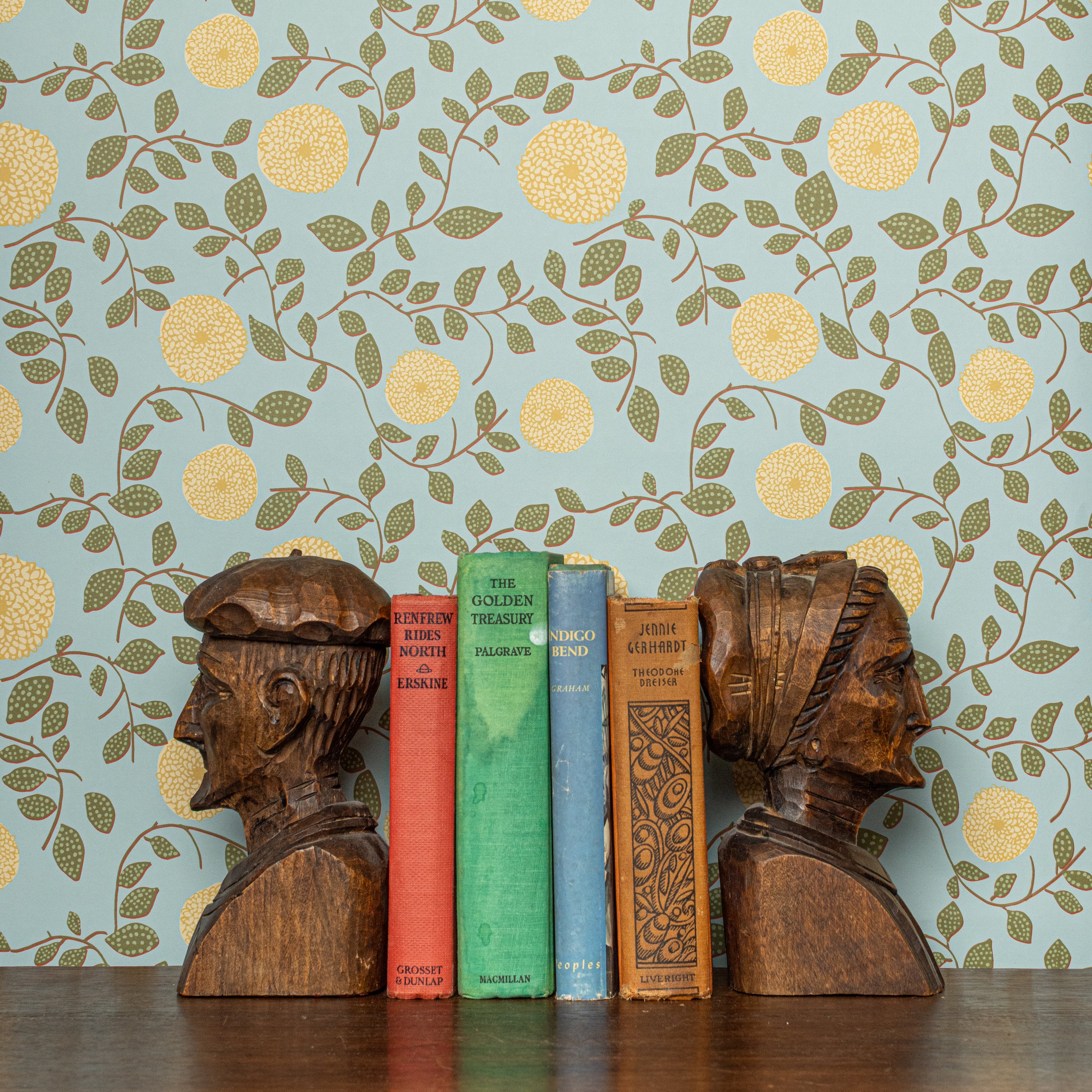 Wooden bookends shaped like human heads on a shelf with books against a floral wallpapered wall.