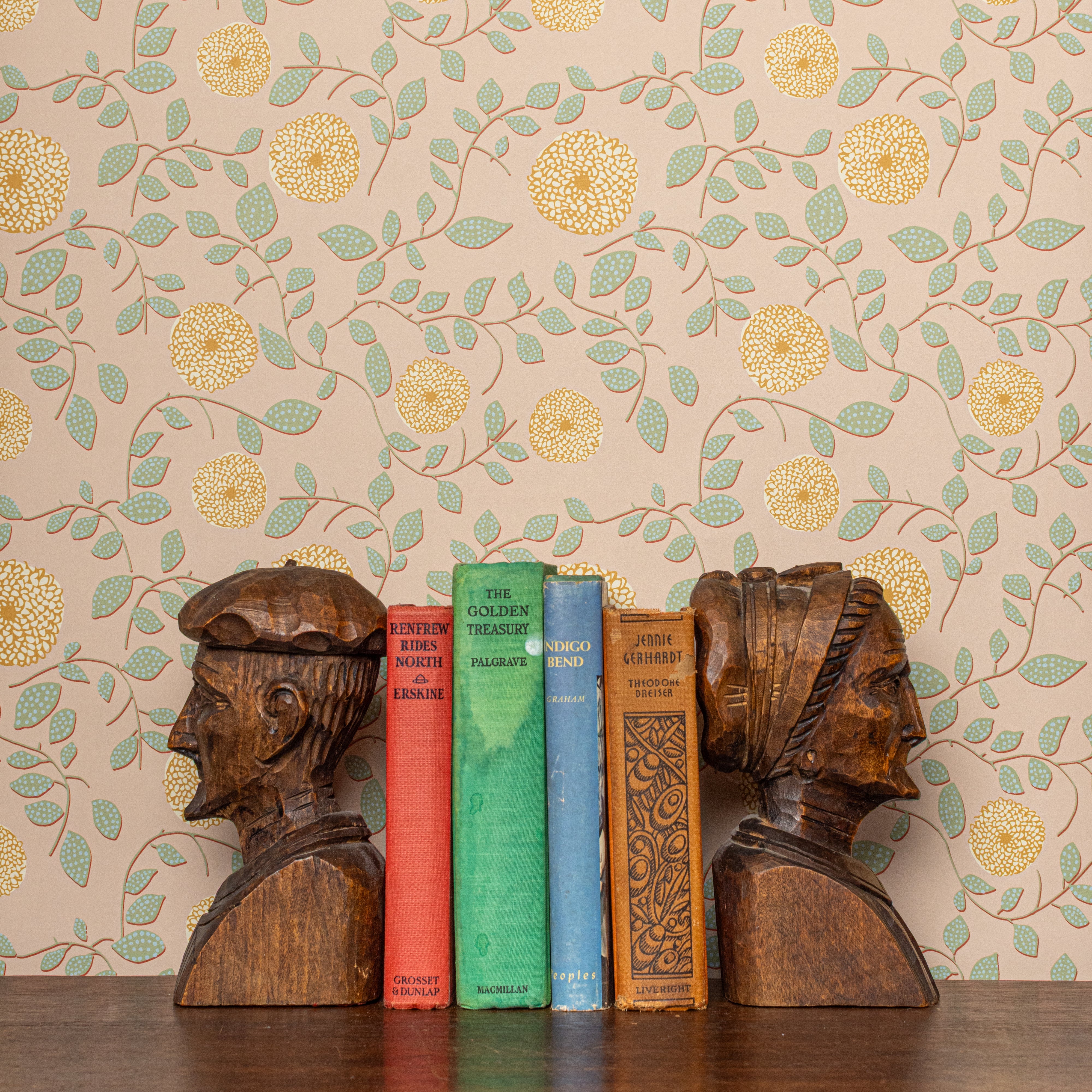 Wooden bookends shaped like human heads with books between them against a floral wallpaper background.