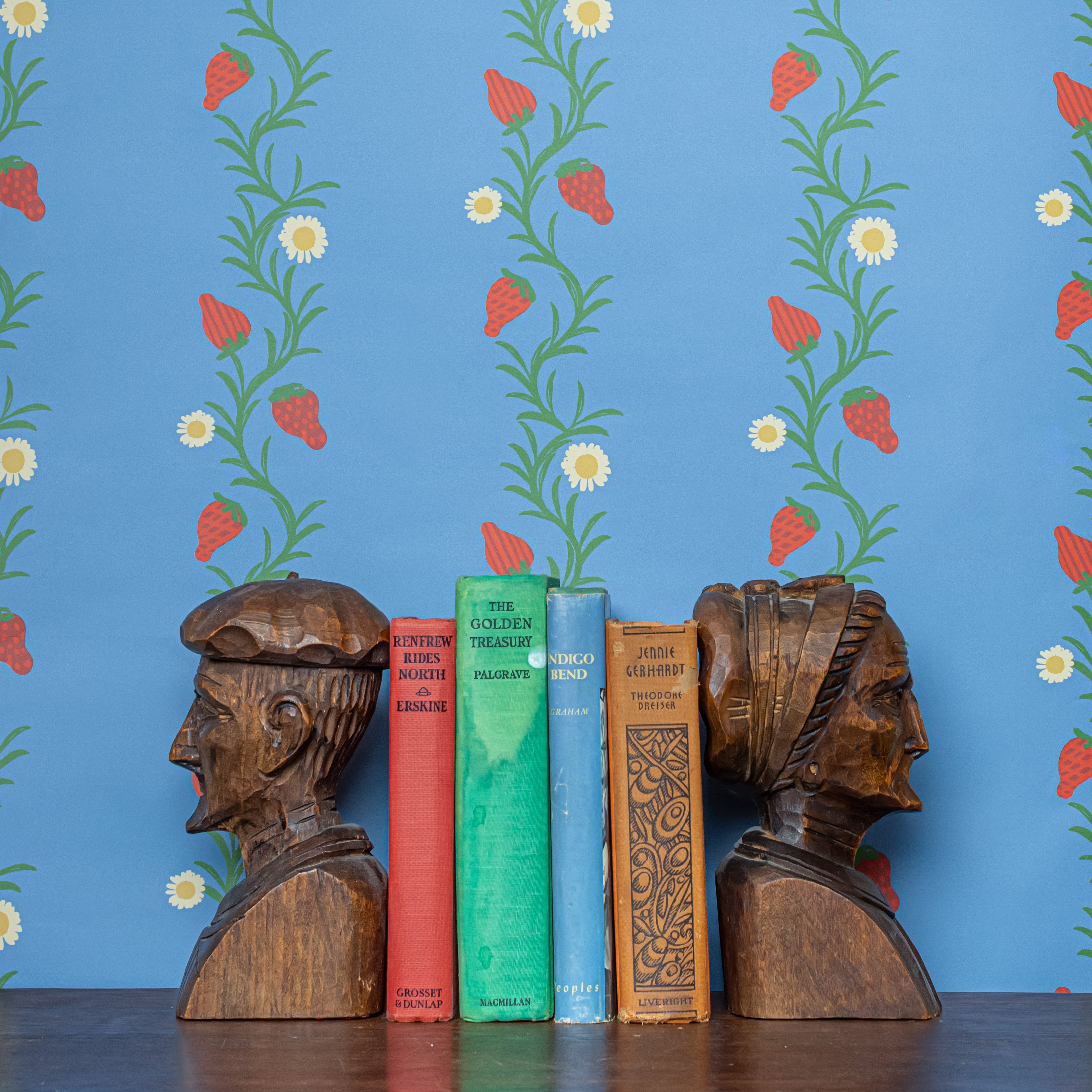 Wooden bookends shaped like faces on a shelf with books against a blue wall paper with strawberry floral pattern