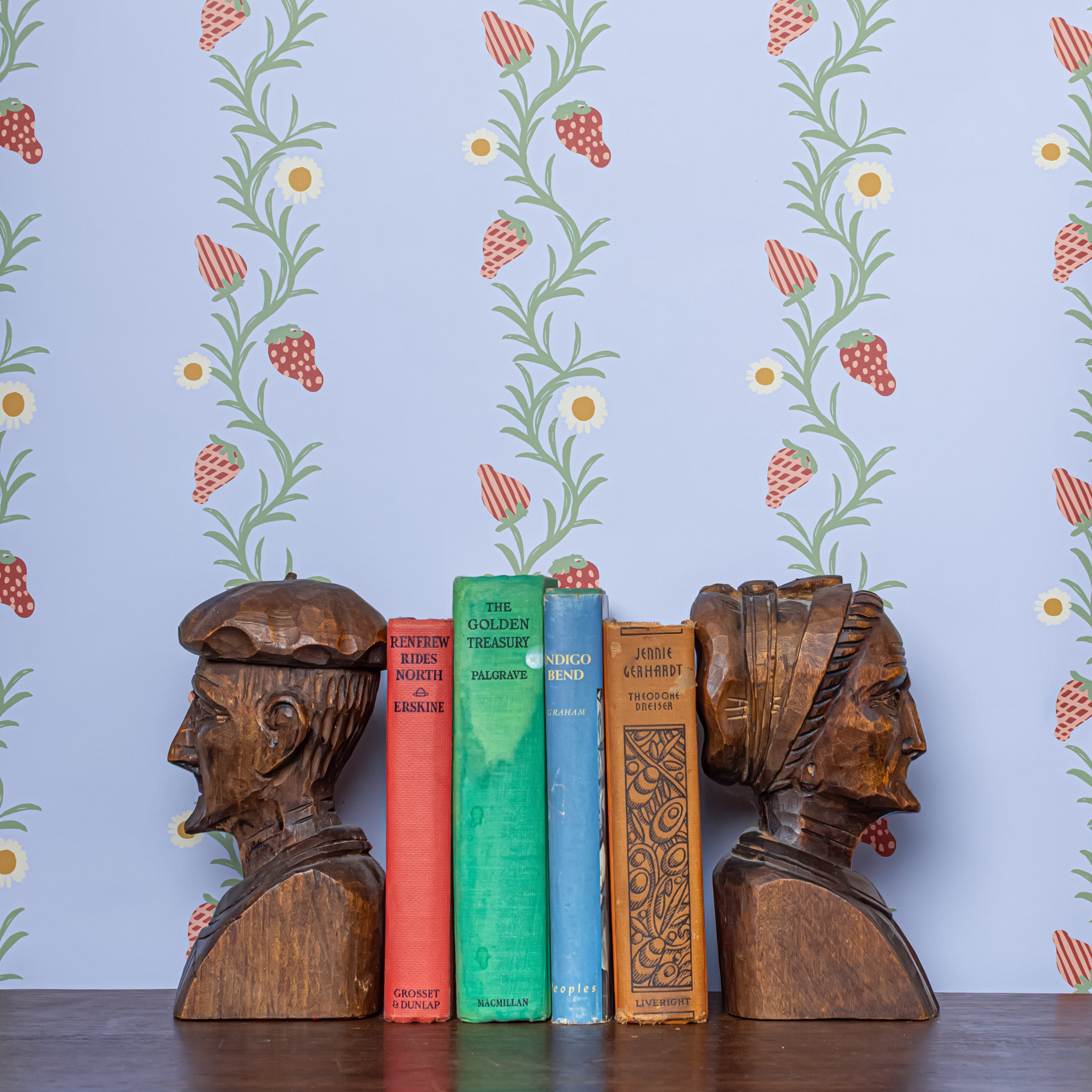 Wooden bookends shaped like human heads on a shelf with books against a strawberry patterned wallpaper in periwinkle.