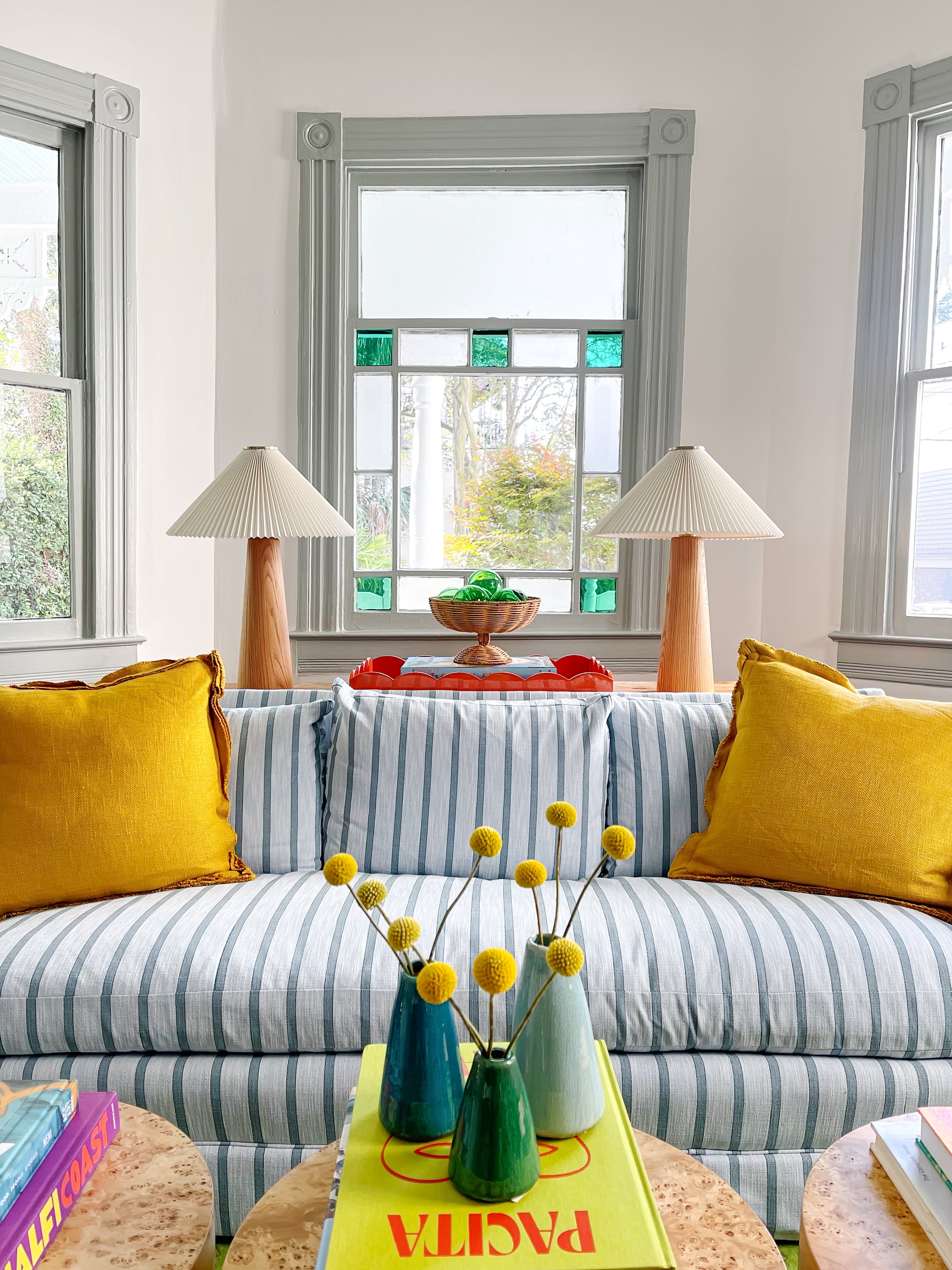 A living room with blue striped furniture, yellow upholstery, and decorative pillows, complemented by a window with a view of autumn leaves and a side table with a lamp and decorative items.