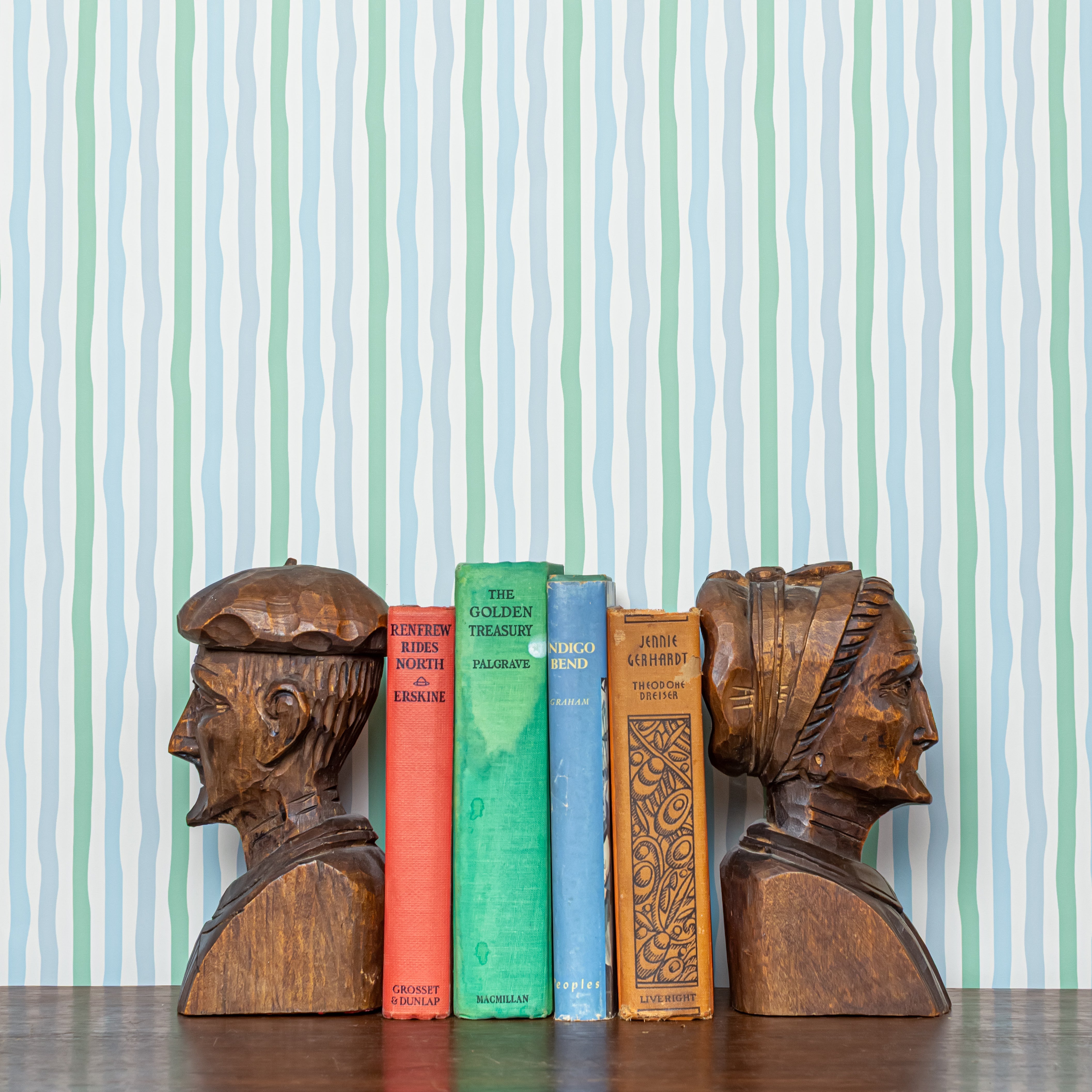 Wooden bookends shaped like human heads on a row of books against a striped  blue and green wallpaper background