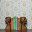 Wooden bookends shaped like faces on a shelf with books against a periwinkle and pink floral wallpaper background