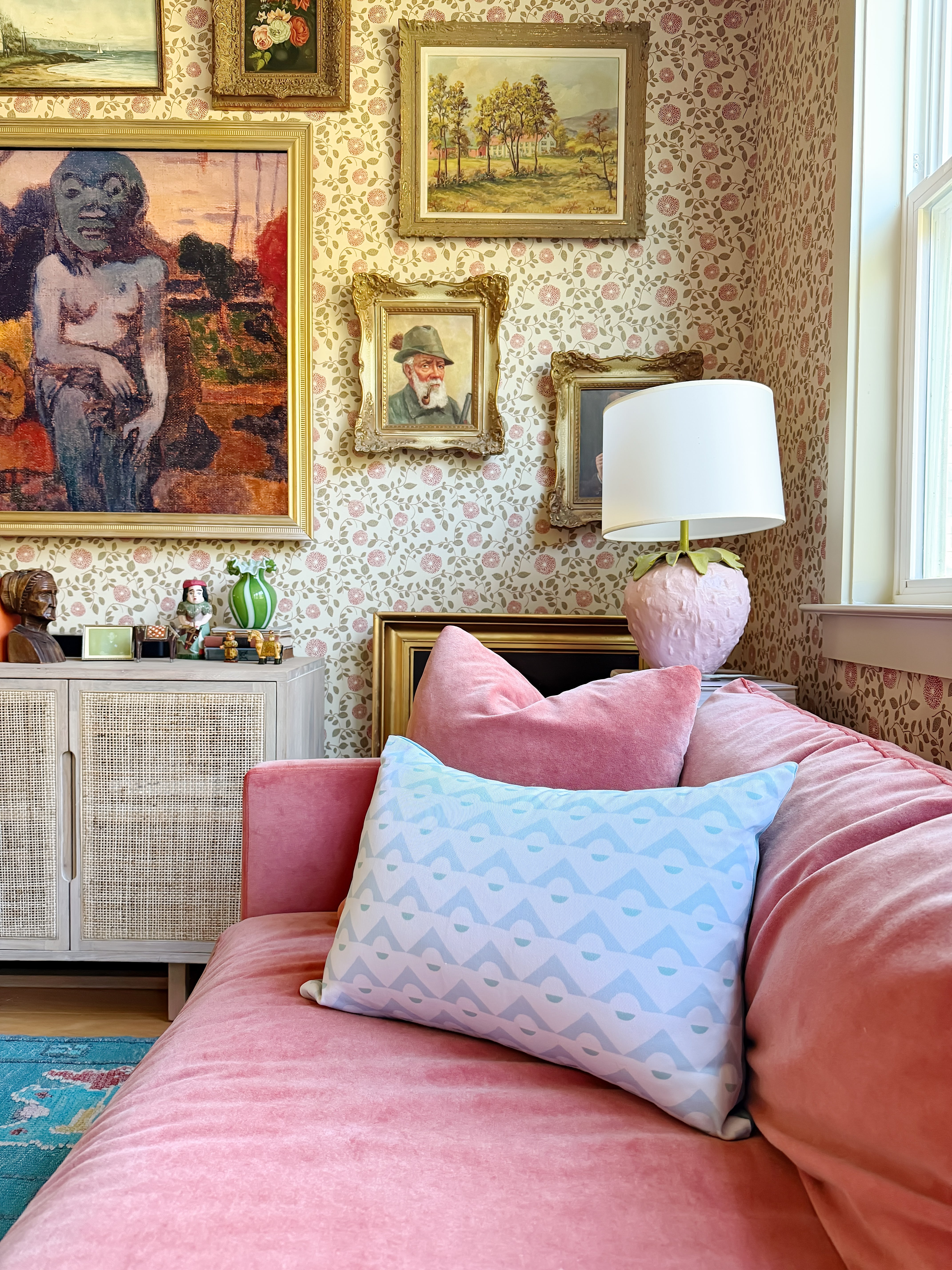 Pink sofa with decorative pillows in a room with floral wallpaper and framed artworks.