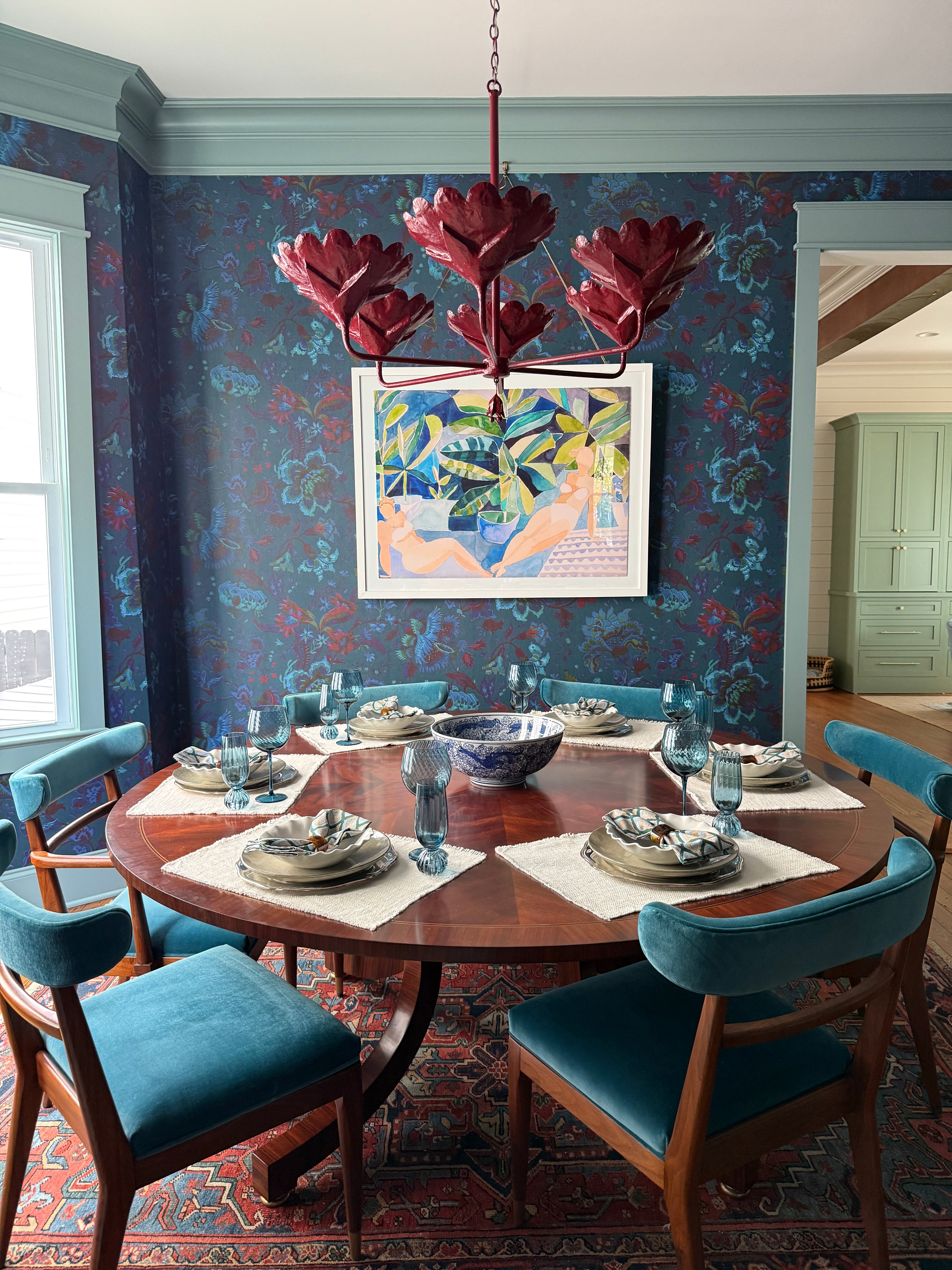 Dining room with a round wooden table, chairs, and a colorful wall painting.
