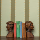 Wooden bookends shaped like faces on a shelf with books against a striped wallpaper in green