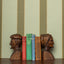Wooden bookends shaped like faces on a shelf with books against a striped wallpaper in green