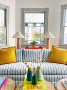 A living room with blue striped furniture, yellow upholstery, and decorative pillows, complemented by a window with a view of autumn leaves and a side table with a lamp and decorative items.