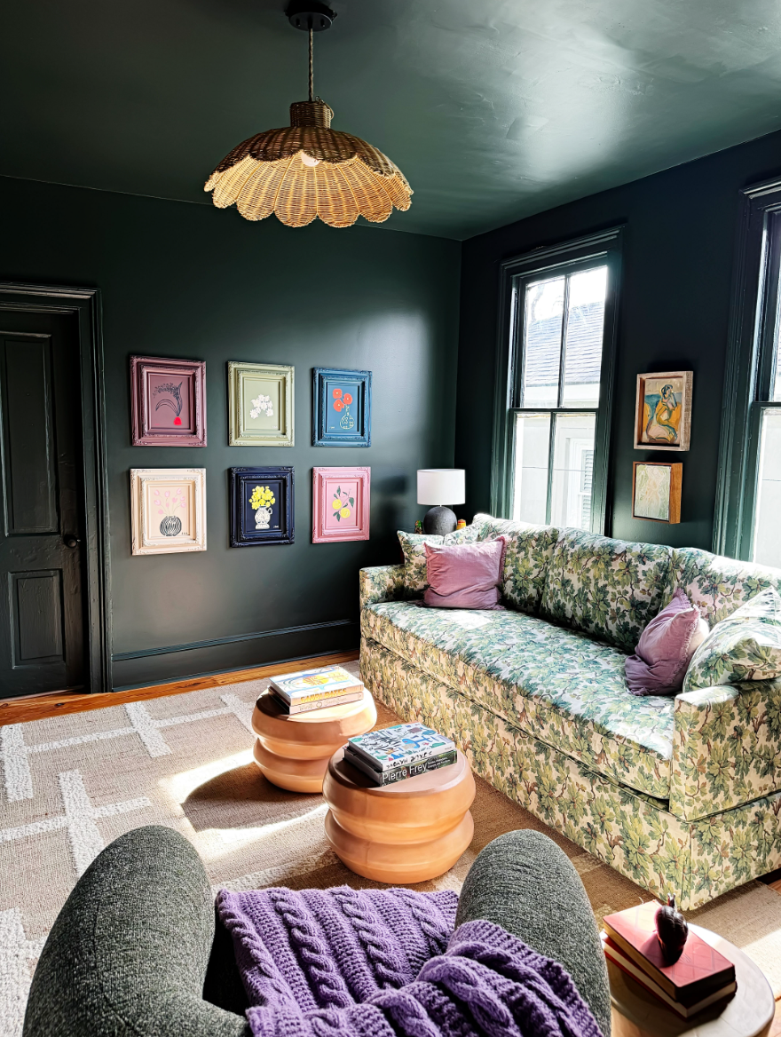 A well-lit den with dark green walls, featuring a hanging ceiling lamp with a conical shade, a floral patterned couch, a round wooden side table with a lamp, and a cozy knit throw on the arm of the couch.